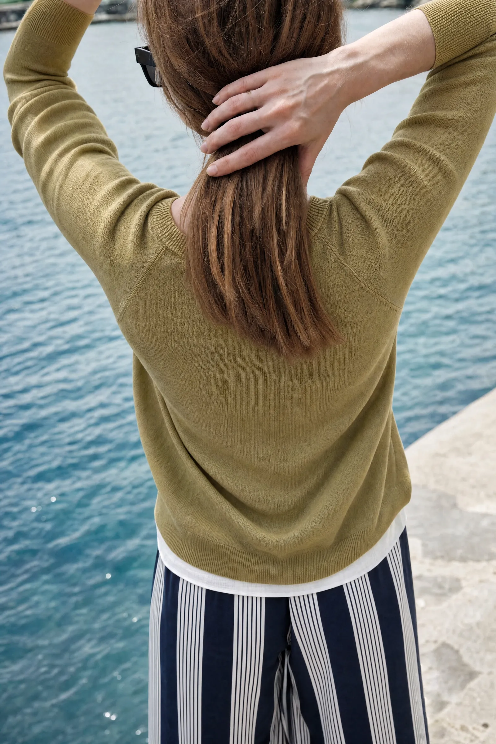 Model seen from behind in an olive sweater and navy striped trousers by the water